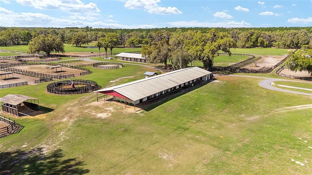 This aerial view showcases a sprawling horse farm with multiple paddocks, a large stable building with a red-trimmed roof, and a secondary building in the background. The property features lush green pastures, mature trees, and a winding driveway, creating a picturesque and well-maintained equestrian estate. The perspective is high-angle, providing a comprehensive view of the entire property.