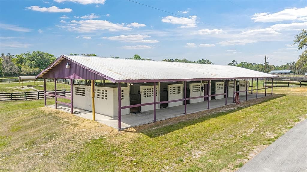 This image showcases a side view of a horse stable or barn structure. The building features multiple stalls with concrete walls and barred openings, all sheltered under a long, metal roof supported by purple-painted posts. The surrounding area is grassy, suggesting a rural or equestrian property.