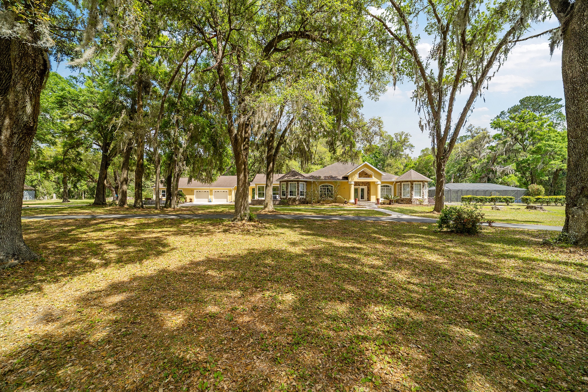 This is a front view of a one-story home with a yellow exterior and a well-manicured lawn. Mature trees with Spanish moss partially obscure the view of the house, adding to the classic southern charm. A circular driveway leads up to the house, suggesting a spacious property.