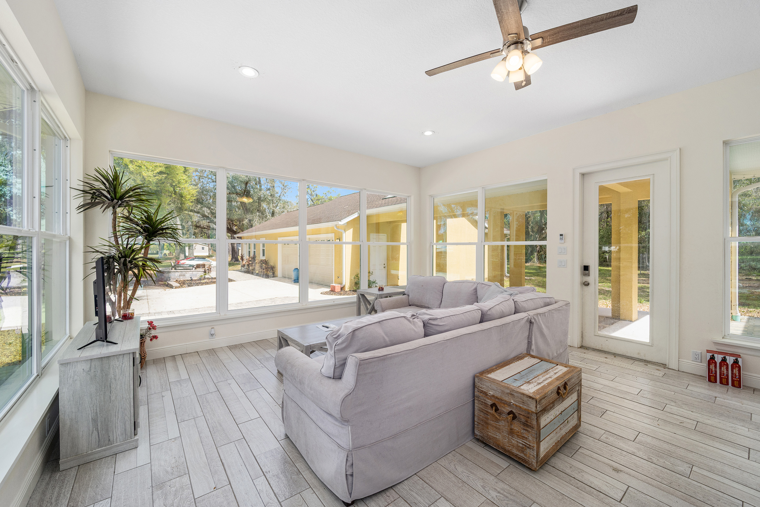 The image showcases a bright and airy sunroom featuring large windows that offer ample natural light and a view of the exterior. A comfortable gray sofa and rustic wooden chest create a relaxed seating area. The light-colored wood-look tile flooring enhances the room's spacious feel, making it an ideal spot for relaxation or entertaining.