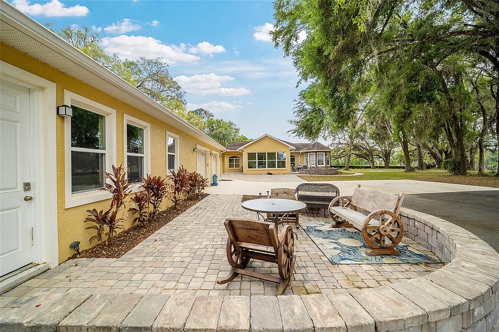 This image showcases a charming patio area with a unique design. The patio features a circular layout with a low brick wall, a stone-paved surface, and rustic wooden furniture, including a rocking chair and a bench with wagon wheel accents. The surrounding landscape includes a well-maintained lawn, mature trees, and a glimpse of the house's exterior, creating a serene and inviting outdoor space.