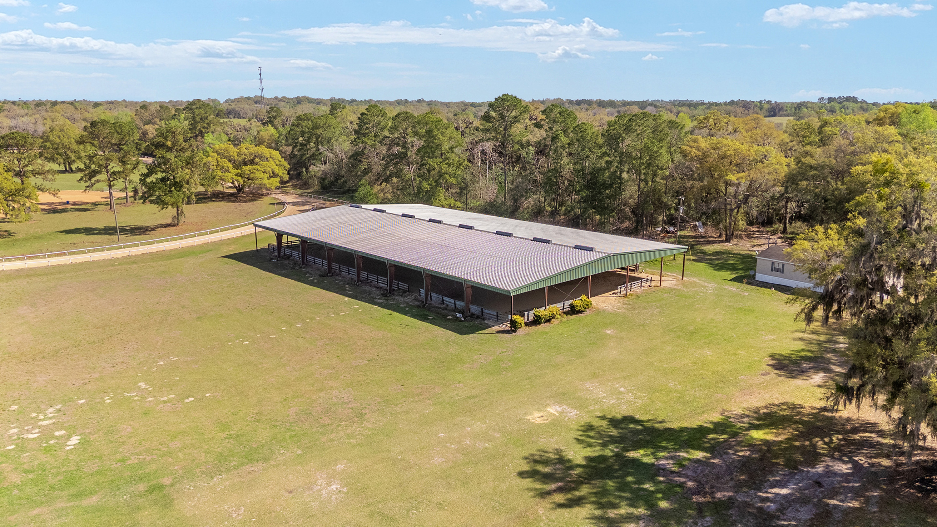 An aerial view showcases a property featuring a large covered structure, possibly a riding arena or stable, situated on a spacious grassy field surrounded by mature trees. A small house is visible near the trees on the right side of the property. The expansive land and rural setting offer privacy and potential for various outdoor activities.