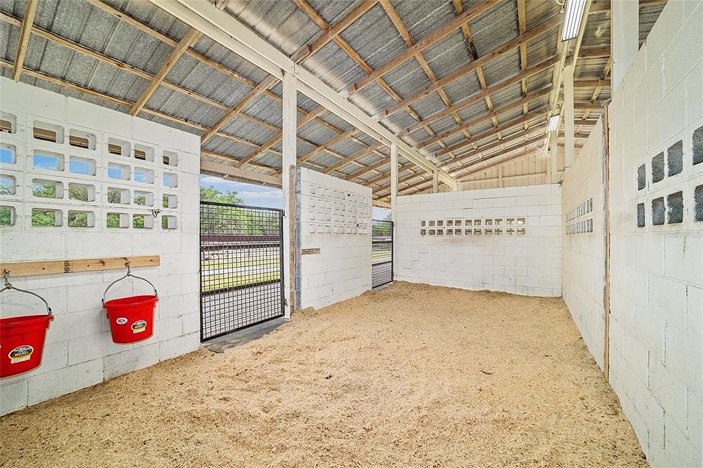 This image showcases a horse stall with white concrete block walls and a metal roof supported by wooden beams. The stall has a sand floor and a metal gate leading to an outdoor area. Two red buckets hang on the wall, adding a pop of color to the otherwise neutral palette.