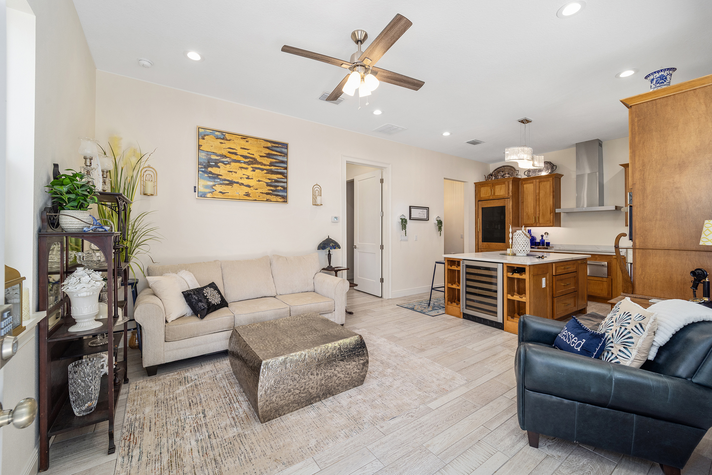 This inviting living room showcases a neutral color palette with a comfortable sofa, a unique metal coffee table, and a dark-toned armchair. An adjacent kitchen area is visible, separated by a bar with a wine cooler. The space is well-lit and appears stylish and comfortable, ideal for relaxing or entertaining.