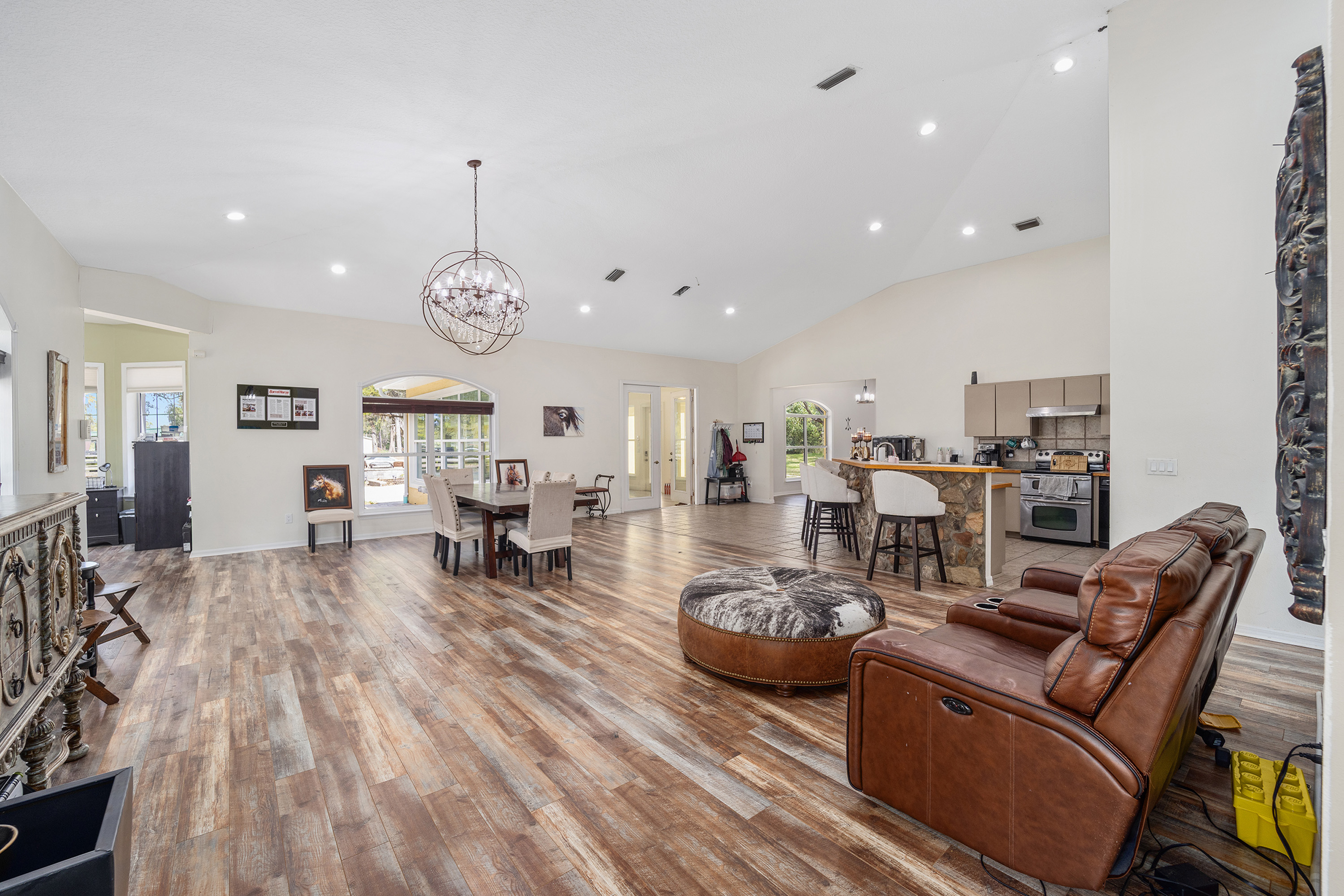 This is a spacious living area with hardwood flooring and vaulted ceilings. It features a dining table, a seating area with a brown leather recliner, and a kitchen bar. The overall feel is open and inviting, lending itself to comfortable living and easy entertaining.