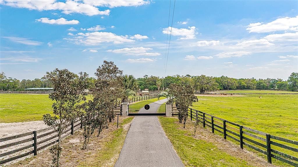 This image showcases the front view of a property, featuring a long driveway leading to an ornate gated entrance. The property is surrounded by green pastures and wooden fencing, suggesting a rural or equestrian estate. The sky is bright blue with scattered clouds, creating a serene and inviting atmosphere.