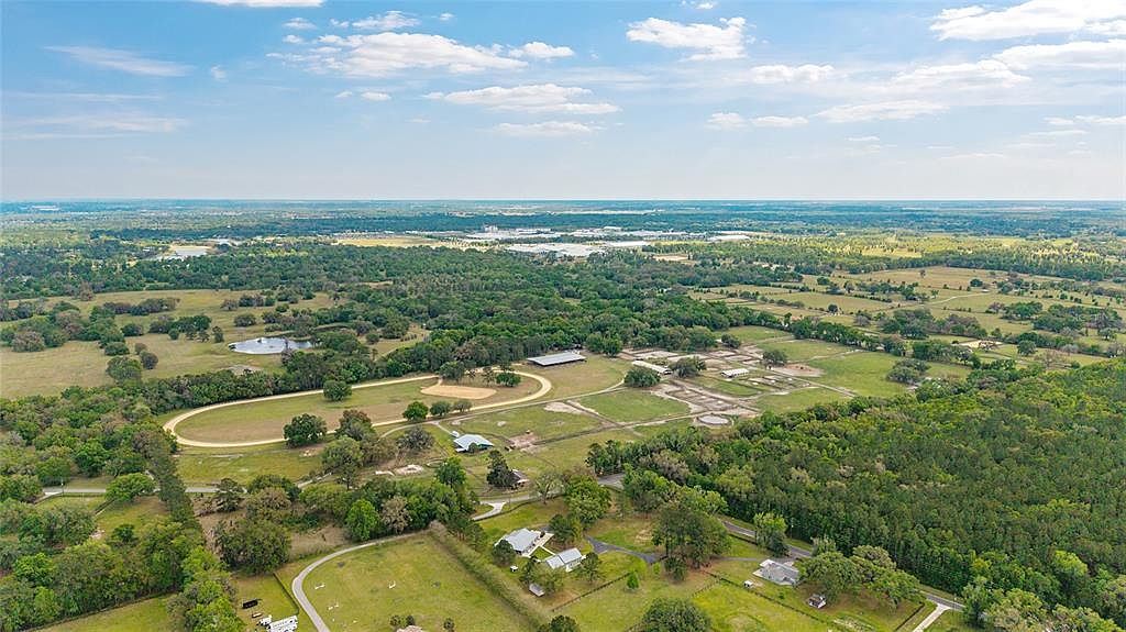 This aerial shot showcases a sprawling estate with a horse track, multiple buildings, and lush green pastures. The property is surrounded by dense trees and features a pond, creating a serene and private setting. The overall impression is one of luxury and spaciousness, ideal for equestrian enthusiasts or those seeking a secluded retreat.