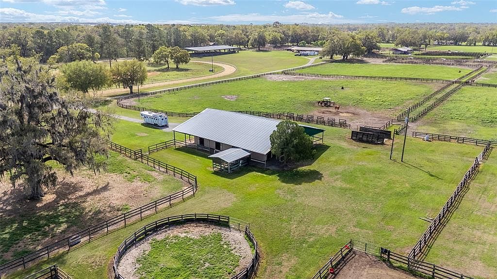This aerial shot showcases a sprawling equestrian estate with lush green pastures, well-maintained fencing, and a central barn structure. A horse trailer is parked near the barn, suggesting an active equestrian lifestyle. The property features multiple paddocks and a riding arena, all set against a backdrop of mature trees and open sky.