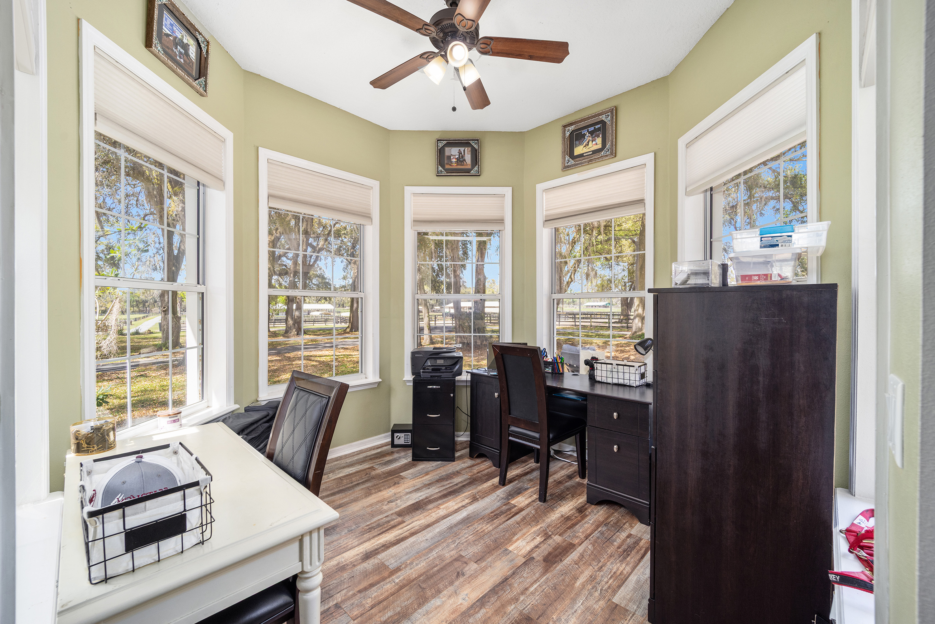 This is an interior shot of a home office featuring a bay window with views of the surrounding property. The room is decorated in neutral tones with wood flooring and includes two desks, a filing cabinet, and a ceiling fan. The space appears well-organized and conducive to work, showcasing a functional and aesthetically pleasing home office.