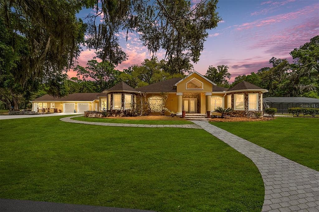 This is a front view of a single-family home with a large, well-manicured lawn and a winding brick pathway leading to the entrance. The house features a combination of stone and stucco exterior, with unique turret-like structures on either side of the main entrance. The property is surrounded by mature trees, creating a sense of privacy and seclusion.