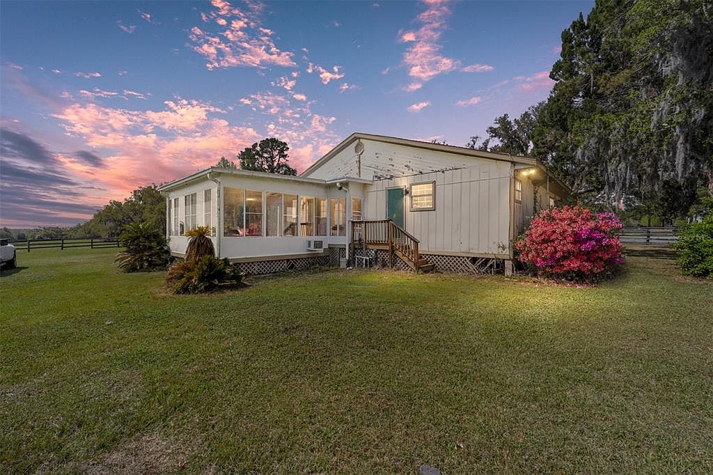 The image showcases the rear exterior of a single-story home with an attached sunroom. The house features light-colored siding, a small deck with stairs, and a well-maintained lawn. A vibrant pink bush adds a pop of color, and mature trees provide a natural backdrop, creating a serene and inviting atmosphere.