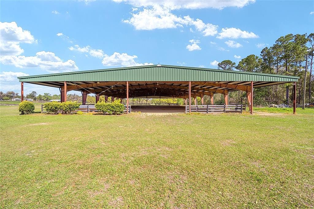 The image showcases a large, open-air riding arena with a green metal roof supported by metal beams. The arena is surrounded by a low wooden fence and lush green grass, creating a spacious and inviting equestrian facility. In the background, trees and a clear blue sky with scattered clouds enhance the rural setting.