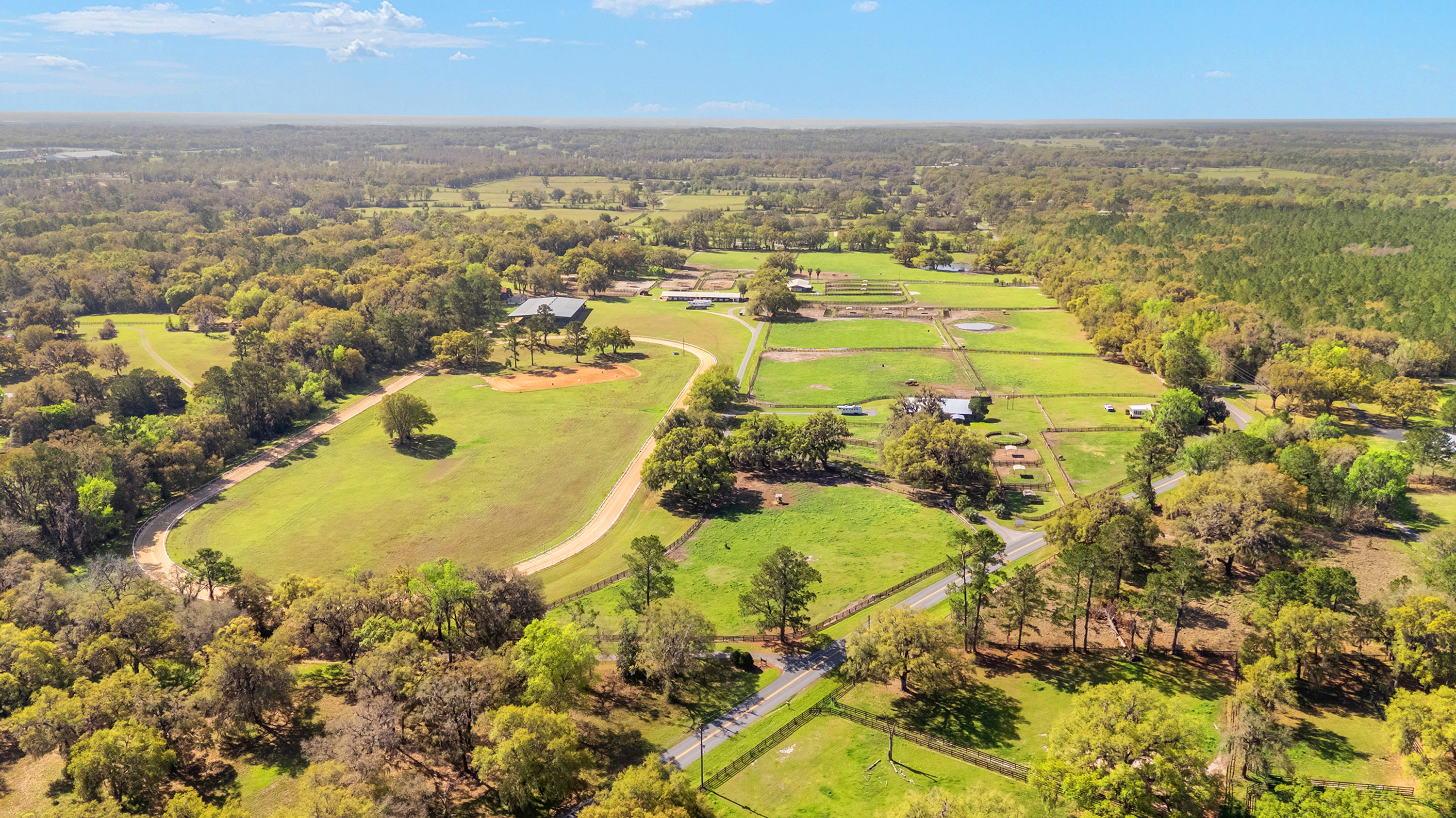 This aerial view showcases a sprawling property with lush green pastures, mature trees, and well-maintained structures. The landscape features riding trails, paddocks, and what appears to be horse riding facilities or a small ranch. The property exudes a sense of privacy and rural charm, perfect for equestrian enthusiasts or those seeking a tranquil country estate.