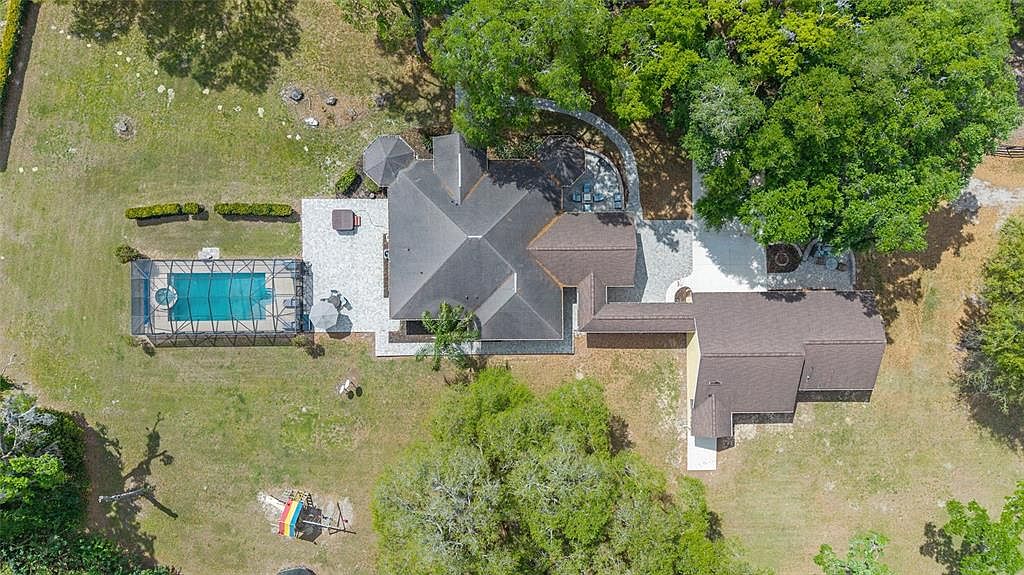 This aerial shot showcases a sprawling property featuring a well-maintained house with a complex roofline, a screened-in pool area, and lush landscaping. The property includes paved patios and walkways, connecting various outdoor spaces, and is surrounded by mature trees and grassy areas, creating a private and serene setting. The overall impression is one of luxury and spaciousness.