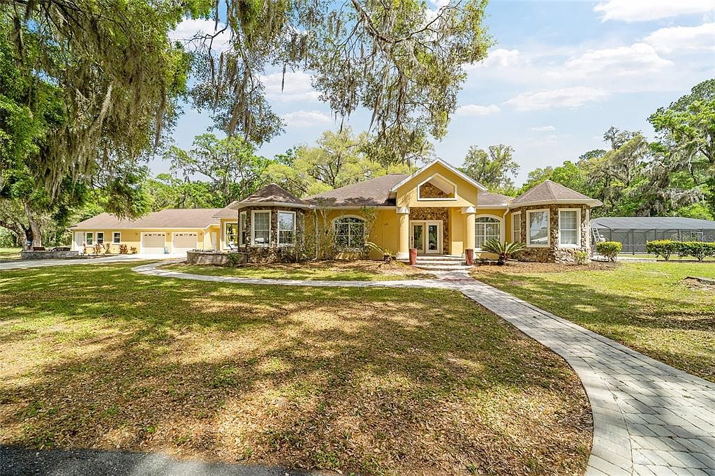 This is a front exterior view of a single-story yellow house with stone accents and a well-manicured lawn. A paved walkway leads to the front entrance, flanked by columns. Mature trees with Spanish moss provide shade, enhancing the property's curb appeal.