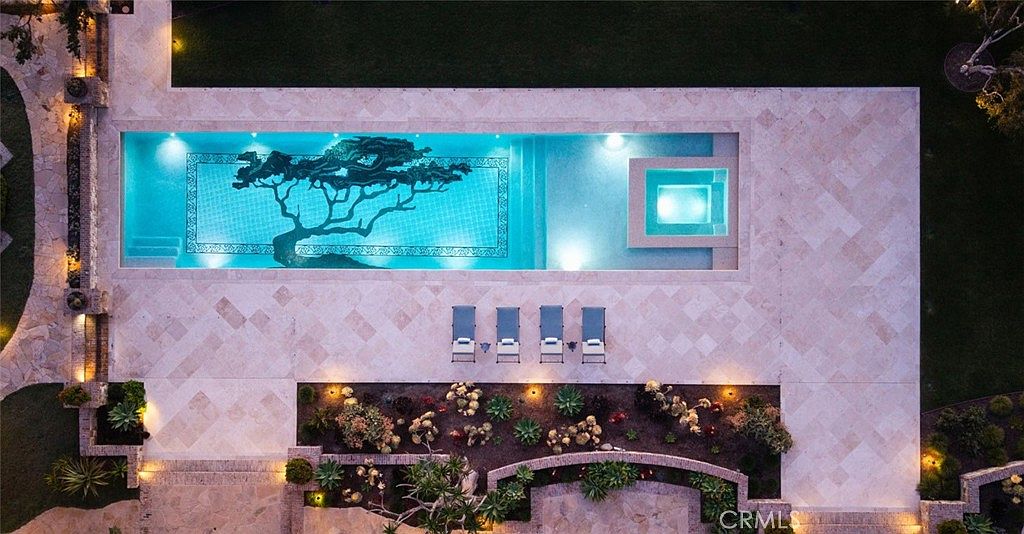 This aerial perspective captures a luxurious rectangular swimming pool featuring a unique, artistic tree mosaic on the pool floor. Adjacent to the pool is a built-in spa, surrounded by expansive light-toned stone decking and four lounge chairs arranged for relaxation. The scene is framed by a landscaped garden area with various desert-style plants and soft, warm accent lighting, creating an inviting and sophisticated outdoor retreat.