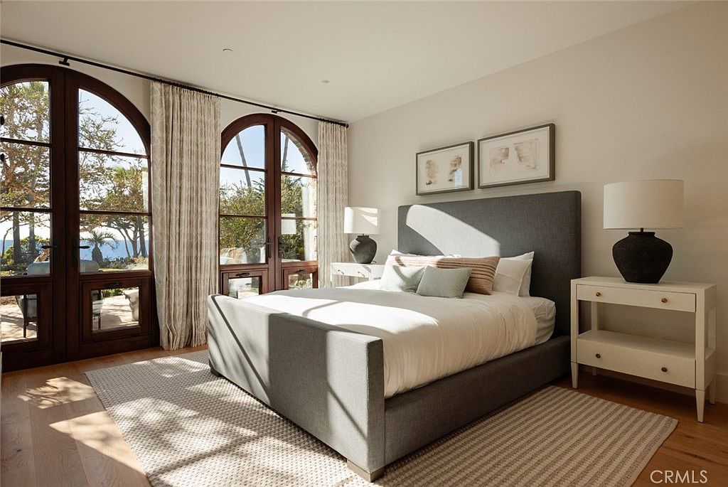 This serene bedroom features a large, upholstered gray bed frame centered against a neutral wall adorned with two framed art pieces. Tall, arched glass doors with dark frames provide abundant natural light and a view of the outdoor landscape, complemented by floor-length curtains. The room is furnished with matching light-toned nightstands topped with dark, textured table lamps, all resting on a light wood floor covered by a textured area rug.
