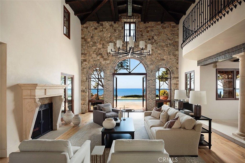 This expansive living room features a dramatic stone-clad chimney breast and a vaulted, exposed-beam ceiling that creates a grand, rustic atmosphere. Large arched glass doors open directly to a stunning ocean view, seamlessly blending indoor and outdoor living spaces. The room is furnished with a plush sofa and armchairs arranged around a dark coffee table, all set upon a neutral area rug over light wood flooring.