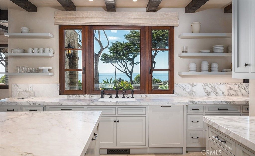 This elegant kitchen features light-toned cabinetry paired with expansive stone countertops and a matching backsplash. A large, multi-pane window centered above the sink provides a stunning, unobstructed view of the ocean and coastal trees. The space is accented by exposed wooden ceiling beams and open shelving, creating a sophisticated, airy atmosphere with a classic coastal aesthetic.
