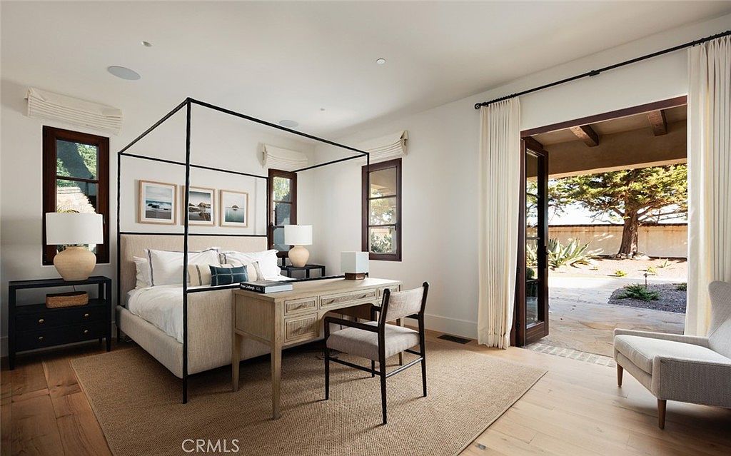 This elegant primary bedroom features a four-poster bed frame and a dedicated desk area, creating a sophisticated multi-functional space. The room is bathed in natural light from large windows and a set of glass doors that open directly onto a private outdoor patio. Neutral tones, light wood flooring, and a textured area rug contribute to a serene and high-end atmosphere.