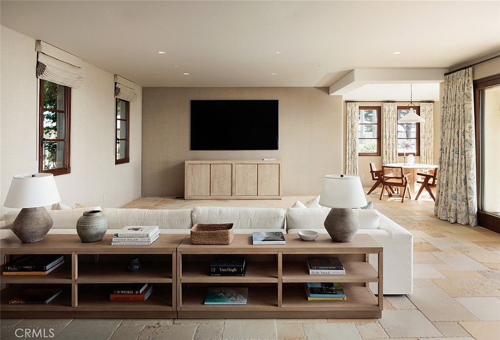 This spacious living room features a neutral color palette with light stone flooring and a large, comfortable sofa positioned in front of a low-profile light wood media console. A wall-mounted television serves as the focal point, while a long, open-shelving console table behind the sofa provides ample display space for books and decor. The room flows seamlessly into an adjacent dining area, creating an open-concept layout filled with natural light from the surrounding windows.