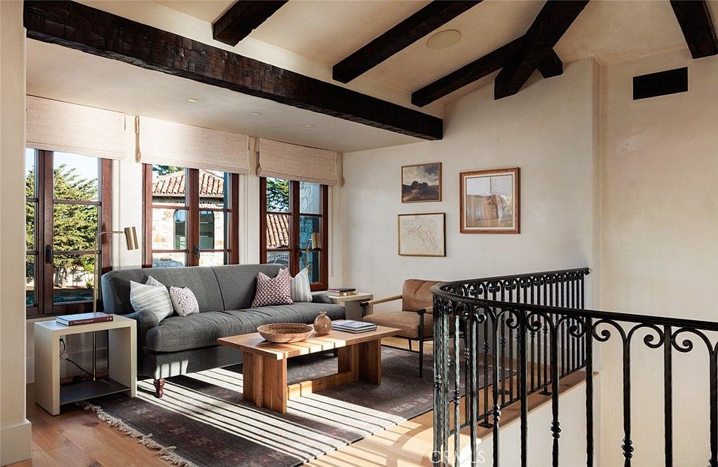 This inviting living room features exposed dark wood ceiling beams and a neutral color palette that creates a warm, sophisticated atmosphere. A plush gray sofa is centered on a patterned area rug, complemented by a light wood coffee table and a leather accent chair. The space is illuminated by natural light streaming through large windows, while an ornate wrought-iron railing overlooks the area, adding a touch of architectural character.