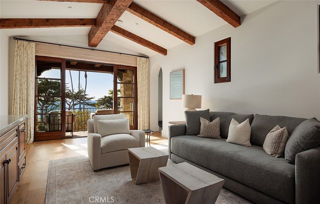 This elegant living room features exposed wood ceiling beams and light-toned hardwood flooring, creating a warm and inviting atmosphere. A plush gray sofa and a matching armchair are arranged around two geometric wooden side tables, while large glass doors open to a balcony with ocean views. The space is bright and airy, blending traditional architectural elements with comfortable, modern furnishings.