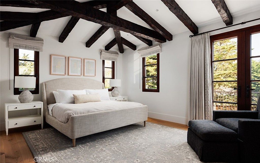 This primary bedroom features a vaulted ceiling with exposed dark wood beams, creating a rustic yet refined aesthetic. The room is anchored by a light-colored upholstered bed, flanked by matching nightstands and lamps, while a large patterned area rug adds texture to the light wood flooring. Large windows and a set of French doors with dark frames allow for abundant natural light, providing a bright and airy atmosphere.