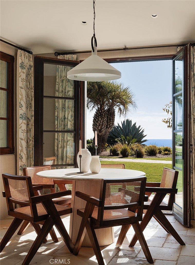 This elegant dining area features a round stone-topped table surrounded by four wooden chairs with woven cane detailing. A modern, minimalist pendant light hangs above the table, which is set with two simple vases and candles. Large, open glass doors provide a seamless transition to a lush outdoor garden with a view of the ocean in the distance.