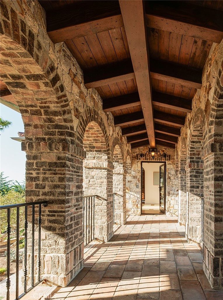 This covered outdoor walkway features rustic brick arches and stone pillars, creating a Mediterranean-inspired aesthetic. The space is topped with a dark wood-beamed ceiling and paved with terracotta-toned tiles, leading toward an open doorway that reveals a bright interior. The perspective looks down the length of the corridor, highlighting the interplay of light and shadow cast by the architectural structure.