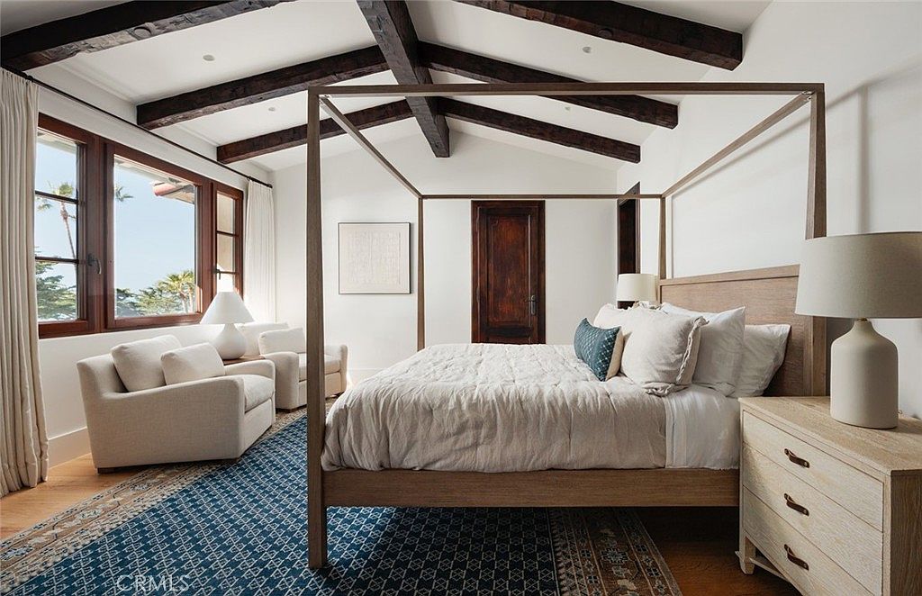 This spacious primary bedroom features a prominent four-poster bed as the central focal point, set against a backdrop of white walls and exposed dark wood ceiling beams. To the left, a cozy seating area with two armchairs is positioned near large windows that offer natural light and views of the outdoors. The room is anchored by a patterned blue rug, and a light wood nightstand with a modern lamp sits beside the bed, creating a serene and sophisticated atmosphere.