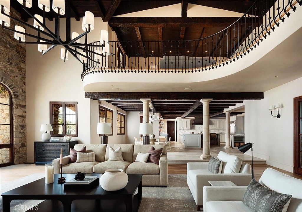 This expansive living room features a dramatic double-height ceiling with exposed dark wood beams and a large, intricate chandelier. The space is anchored by a neutral-toned sofa and armchairs, with a stone-clad wall on the left and a mezzanine balcony overlooking the area. The open-concept layout flows seamlessly into a kitchen area in the background, defined by stone columns and a warm, sophisticated aesthetic.