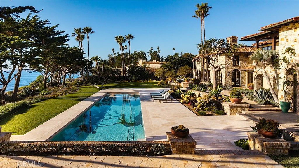 This expansive outdoor space features a long, rectangular swimming pool set against a backdrop of the ocean and a lush, manicured lawn. The stone-clad residence showcases Mediterranean-inspired architecture with warm-toned masonry and a tiled roof, complemented by a spacious stone patio furnished with lounge chairs. The scene is framed by mature trees and tropical landscaping, creating a serene and luxurious coastal atmosphere.