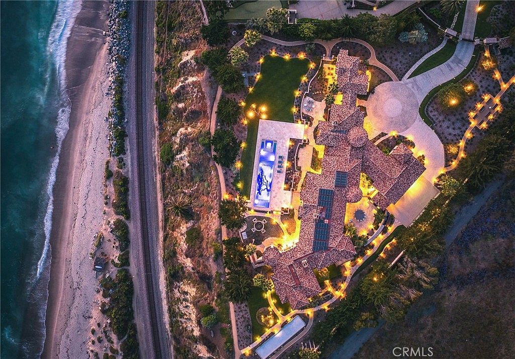 This stunning aerial perspective captures a sprawling luxury estate situated on a coastal cliff overlooking the ocean. The property features an expansive multi-winged residence with a tiled roof, a large rectangular swimming pool, and meticulously landscaped grounds illuminated by warm evening lighting. A private railway line runs along the beach below, emphasizing the dramatic and secluded nature of this prime waterfront location.