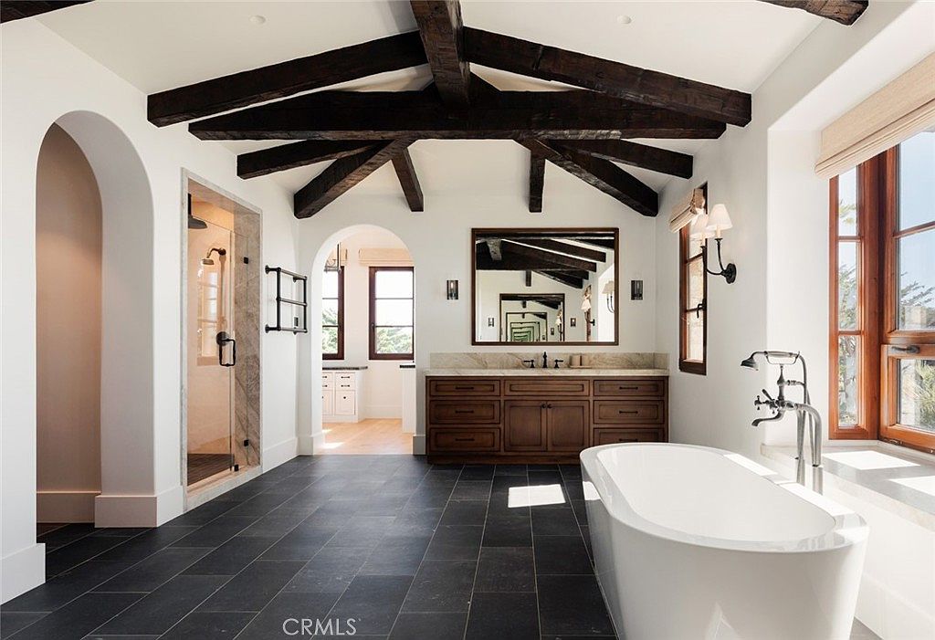 This spacious primary bathroom features a striking vaulted ceiling with exposed dark wood beams and a large, freestanding soaking tub positioned near a window. A double vanity with a stone countertop and a large mirror sits opposite a walk-in glass-enclosed shower, all set against dark slate-style floor tiles. The room is bright and airy, with an arched doorway leading to an adjacent space, creating a sophisticated and luxurious atmosphere.
