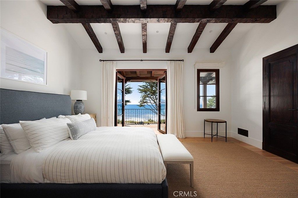 This elegant primary bedroom features exposed dark wood ceiling beams and a neutral color palette that highlights the ocean view through the open French doors. A plush bed with striped bedding sits centered in the room, complemented by a minimalist bench at the foot and a textured area rug. The space is framed by white walls and a dark wood door, creating a sophisticated coastal aesthetic.