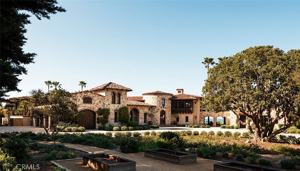 This expansive Mediterranean-style estate features a multi-gabled roofline with terracotta tiles and a facade constructed from textured stone. The front elevation showcases a grand arched entryway, a prominent circular tower element, and a large wooden garage door, all set against a meticulously landscaped courtyard with gravel paths and raised garden beds. A large, mature tree frames the right side of the composition, providing a sense of scale and established elegance to the property.