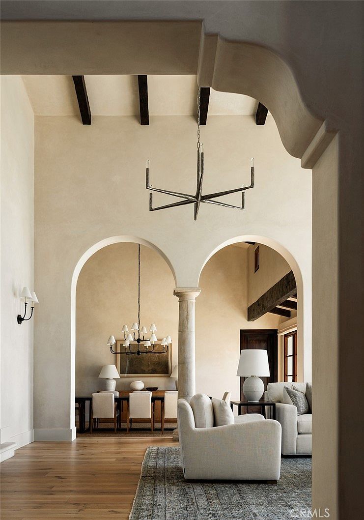 This elegant interior view captures a sophisticated open-concept living and dining area defined by dramatic arched openings and exposed ceiling beams. The space features a neutral color palette, light wood flooring, and a textured area rug, creating a serene and high-end atmosphere. A modern, minimalist chandelier hangs in the foreground, while a secondary dining area is visible through the central archway, showcasing a cohesive design aesthetic.