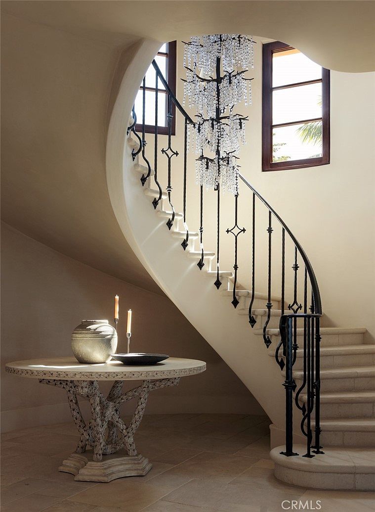 This elegant foyer features a grand curved staircase with intricate wrought-iron railings and stone steps. A large, ornate crystal chandelier hangs from the ceiling, casting a sophisticated glow over the space, while a round pedestal table with candles sits in the foreground. The neutral-toned walls and stone flooring create a timeless, luxurious atmosphere.