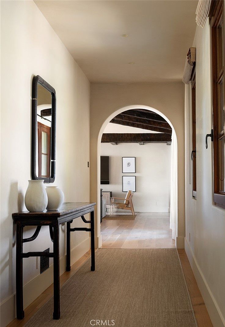 This elegant hallway features a neutral color palette and light wood flooring, leading through a graceful arched doorway into an adjacent living space. A dark, slender console table topped with two white vases sits beneath a rectangular mirror, creating a sophisticated focal point. The perspective is centered, drawing the eye through the archway to reveal a glimpse of a bright room with exposed wooden ceiling beams and framed artwork.