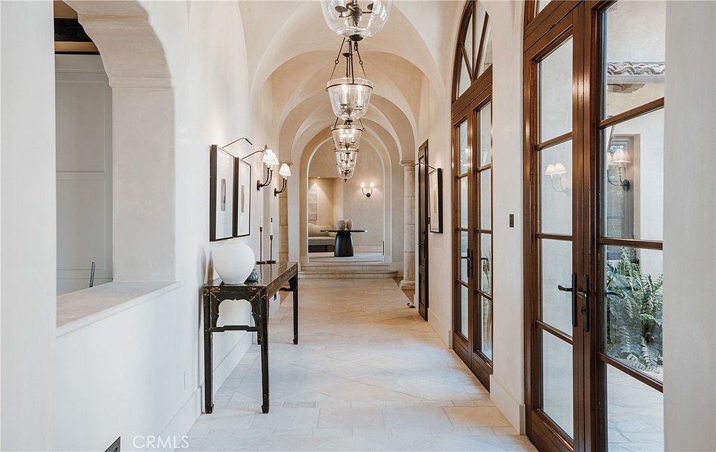 This elegant, arched hallway features a series of glass-paned doors on the right and a long, narrow console table against the left wall. The space is illuminated by a sequence of hanging glass lanterns suspended from the vaulted ceiling, creating a rhythmic and sophisticated atmosphere. The light-toned stone flooring and neutral walls contribute to a bright, airy, and luxurious aesthetic.