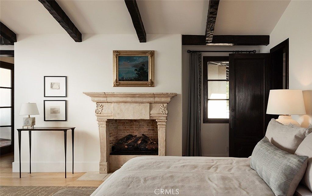This elegant primary bedroom features a prominent stone fireplace with a detailed mantel as the central focal point, topped by a framed landscape painting. The room is characterized by exposed dark wood ceiling beams, white walls, and a neutral-toned bed with textured bedding. A slim console table with a lamp sits to the left, while a dark wood door and a window with floor-length curtains add depth to the space.