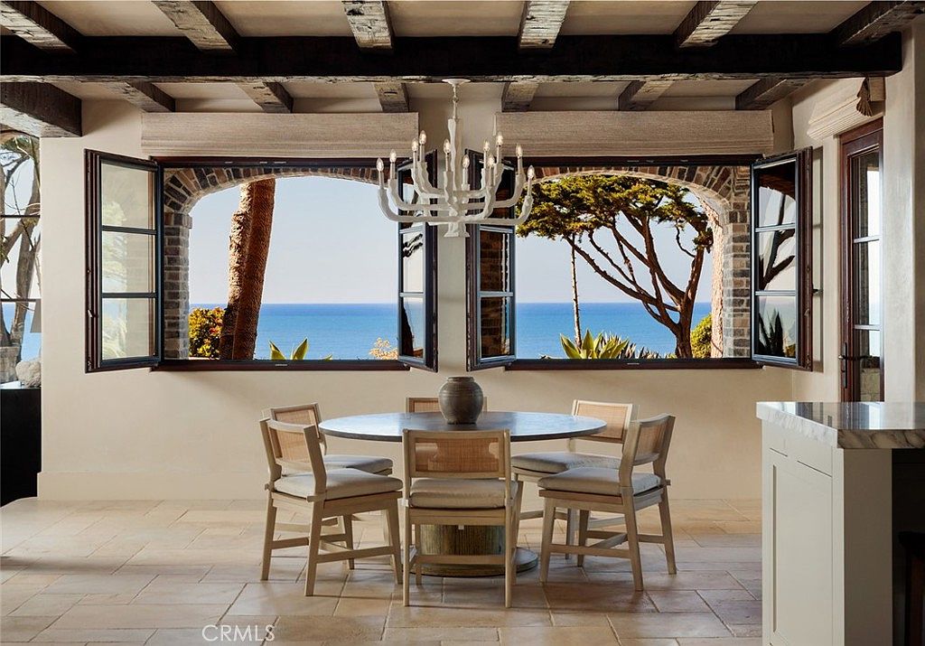 This elegant dining room features a circular wooden table surrounded by four light-toned chairs with woven backrests, positioned beneath a white chandelier. The space is defined by exposed dark wood ceiling beams and light-colored stone flooring, creating a rustic yet refined atmosphere. Large, open casement windows frame a stunning, unobstructed view of the blue ocean and coastal trees, serving as the room's primary focal point.