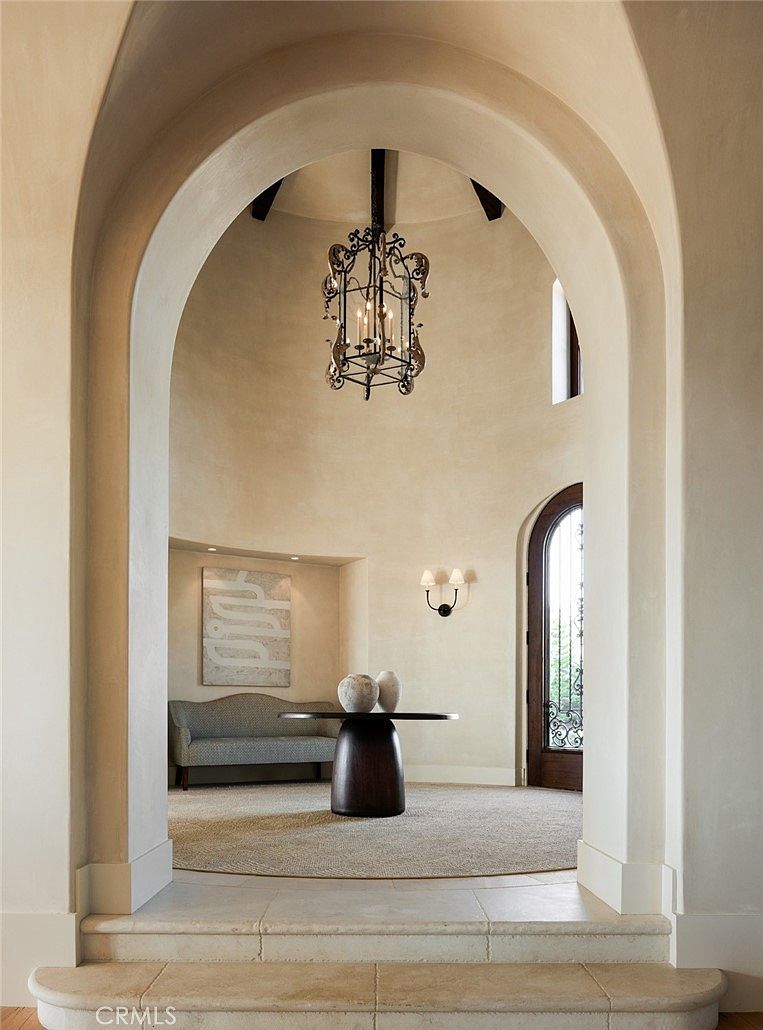 This elegant foyer features a dramatic rotunda design with a high, vaulted ceiling and a prominent wrought-iron chandelier. A central pedestal table sits atop a circular rug, framed by a curved wall niche displaying artwork and a sophisticated settee. The perspective looks through a grand arched stone doorway, highlighting the transition from the tiled steps into the refined, neutral-toned circular hall.