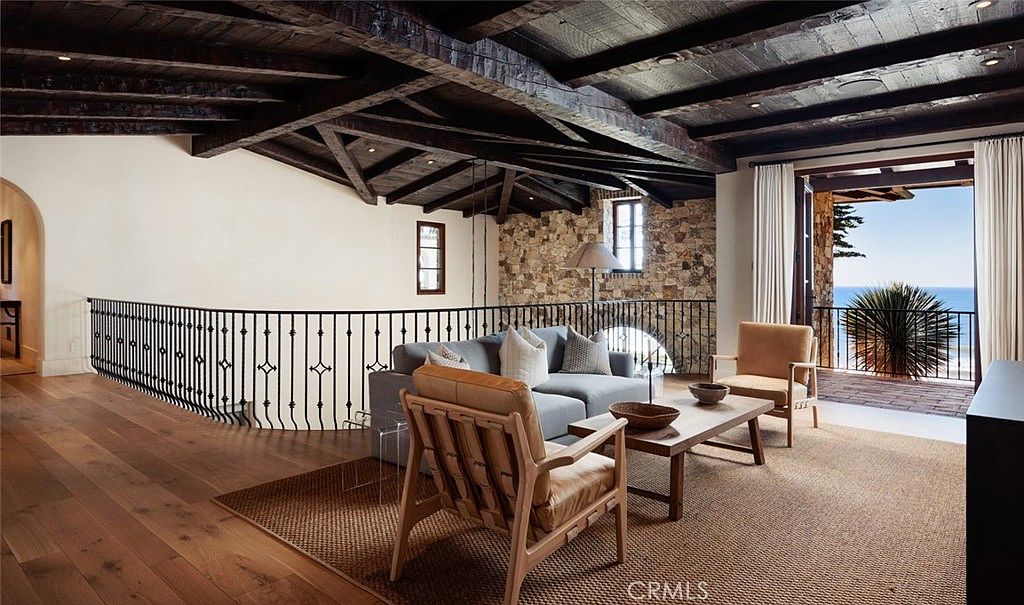 This elegant living area features a dramatic vaulted ceiling with exposed dark wood beams and a rustic stone wall. A comfortable seating arrangement is centered on a textured rug, while a wrought-iron railing overlooks the space below. Large glass doors open to a balcony, providing a seamless transition to the outdoor view and natural light.