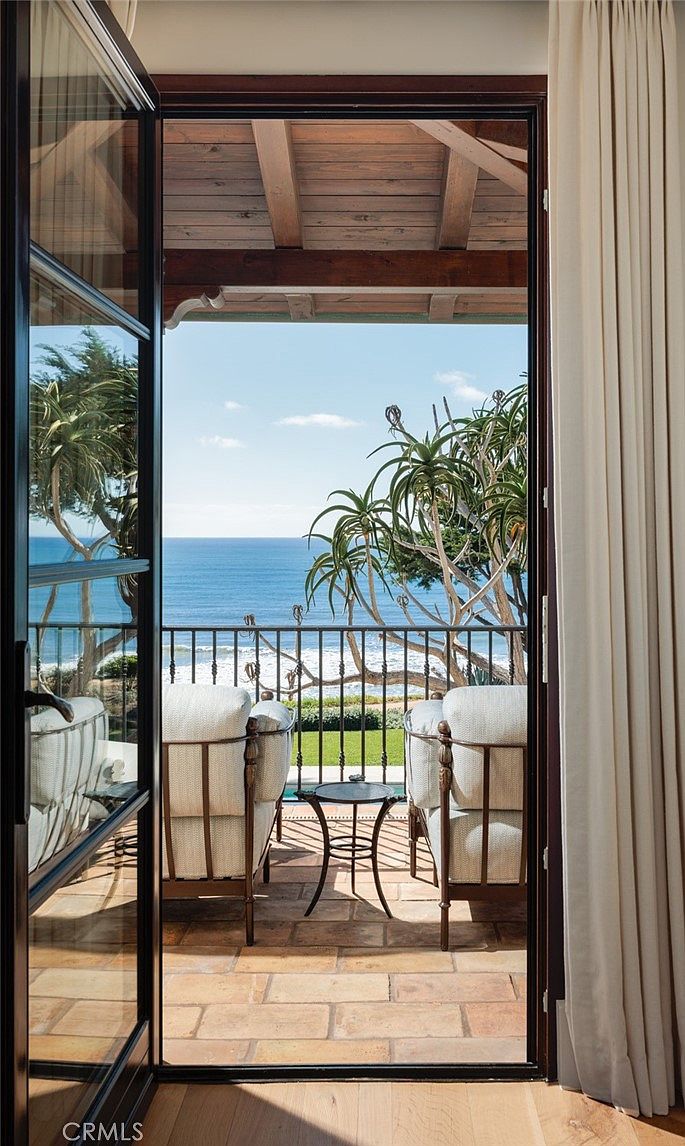 This view from an interior doorway captures a serene balcony overlooking the ocean. The space is furnished with two comfortable armchairs and a small side table, set against a backdrop of lush greenery and a clear blue sky. The stone-tiled floor and wrought-iron railing create a classic, inviting atmosphere perfect for enjoying the coastal scenery.