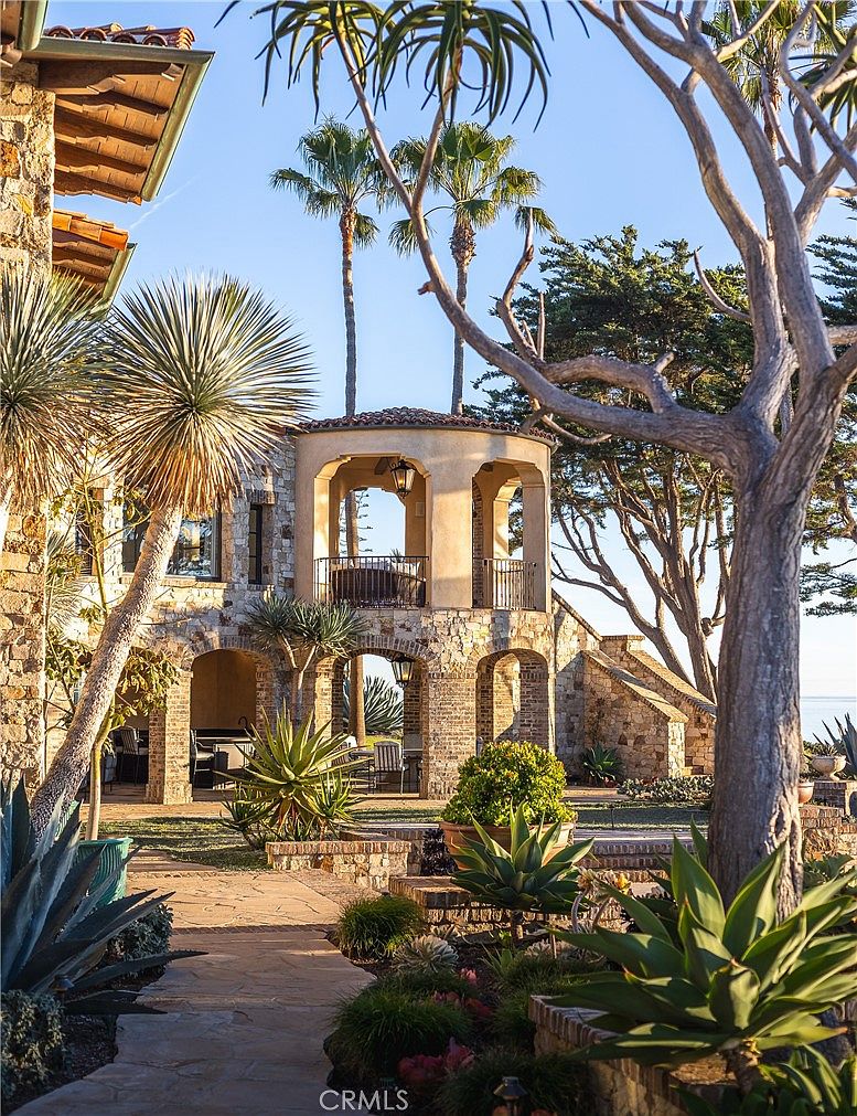 This Mediterranean-style estate features a multi-level stone facade with arched loggias and a prominent second-story balcony overlooking a lush, landscaped garden. The scene is captured from a low angle, emphasizing the textured stone masonry, mature palm trees, and the intricate stone pathway leading toward the covered outdoor living area. The warm, golden hour lighting enhances the rustic charm and coastal elegance of the property's exterior architecture.