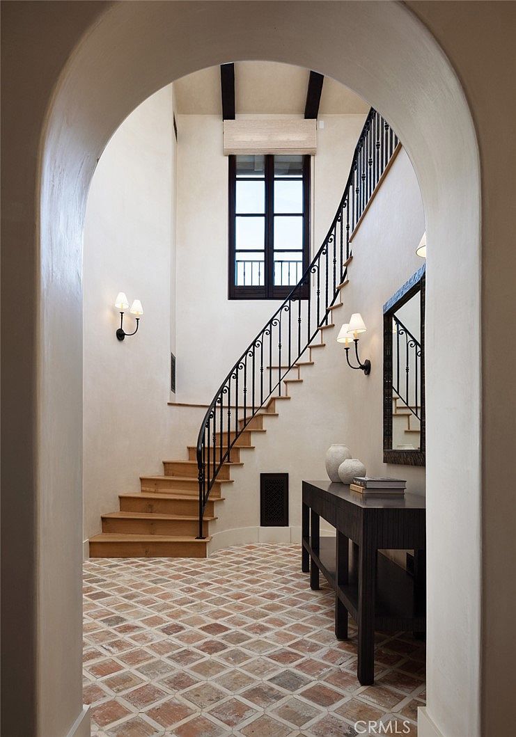 This elegant entryway features a curved staircase with a dark metal railing and light wood treads, set against smooth, neutral-toned walls. The floor is covered in a classic terracotta-style patterned tile, and a dark wood console table sits beneath a framed mirror. The perspective is framed through a large, arched doorway, highlighting the tall window at the top of the stairs and the warm, ambient glow from wall-mounted sconces.
