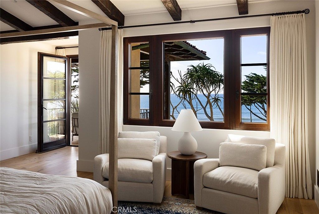 This serene bedroom features a four-poster bed frame and a cozy seating area with two plush armchairs flanking a small round side table. Large windows provide a stunning, unobstructed view of the ocean and coastal landscape, filling the room with natural light. The space is decorated in a neutral, calming palette with light wood flooring and exposed dark wood ceiling beams, creating an elegant and tranquil atmosphere.