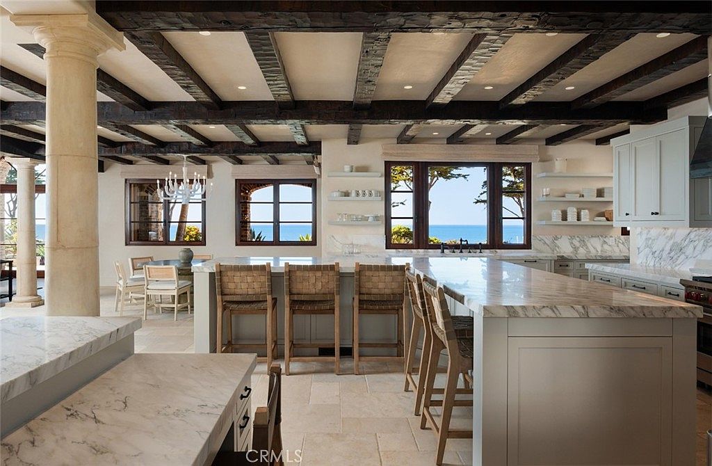 This spacious, open-concept kitchen features a large central island with stone countertops and woven bar stools, perfect for casual dining. The room is defined by dark, exposed wooden ceiling beams and light-toned stone flooring, creating a sophisticated coastal aesthetic. Large windows frame a stunning ocean view, while white cabinetry and open shelving provide a bright, airy feel to the space.